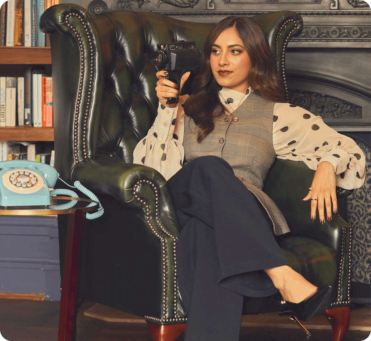 A woman sits in a green leather armchair holding a vintage camera, with a blue rotary phone on a table beside her and bookshelves in the background.