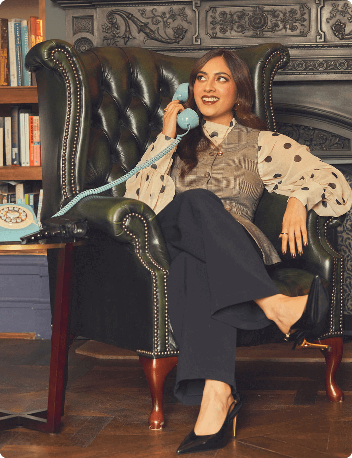 A woman sits in a green leather armchair, smiling while talking on a blue rotary phone. She wears a polka-dot blouse, vest, dark pants, and black heels, with a bookshelf in the background.
