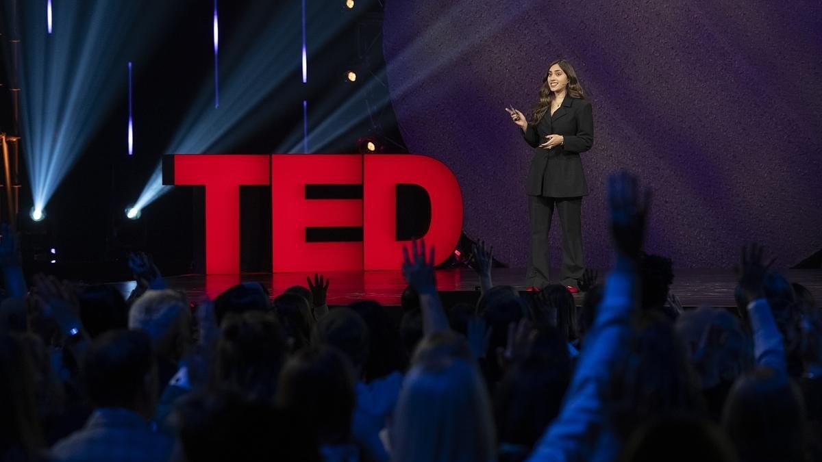 A woman in a black suit stands on stage next to large red TED letters, speaking to an audience with raised hands under stage lights.