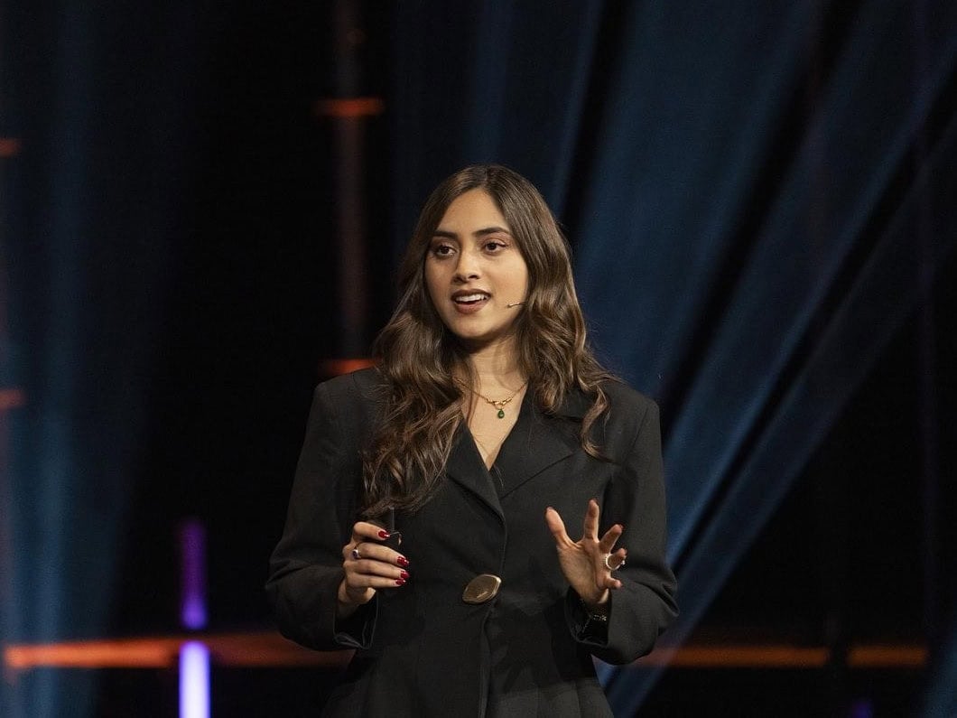 A woman in a black blazer speaks on stage, gesturing with her hands, with dark curtains and stage lights in the background.