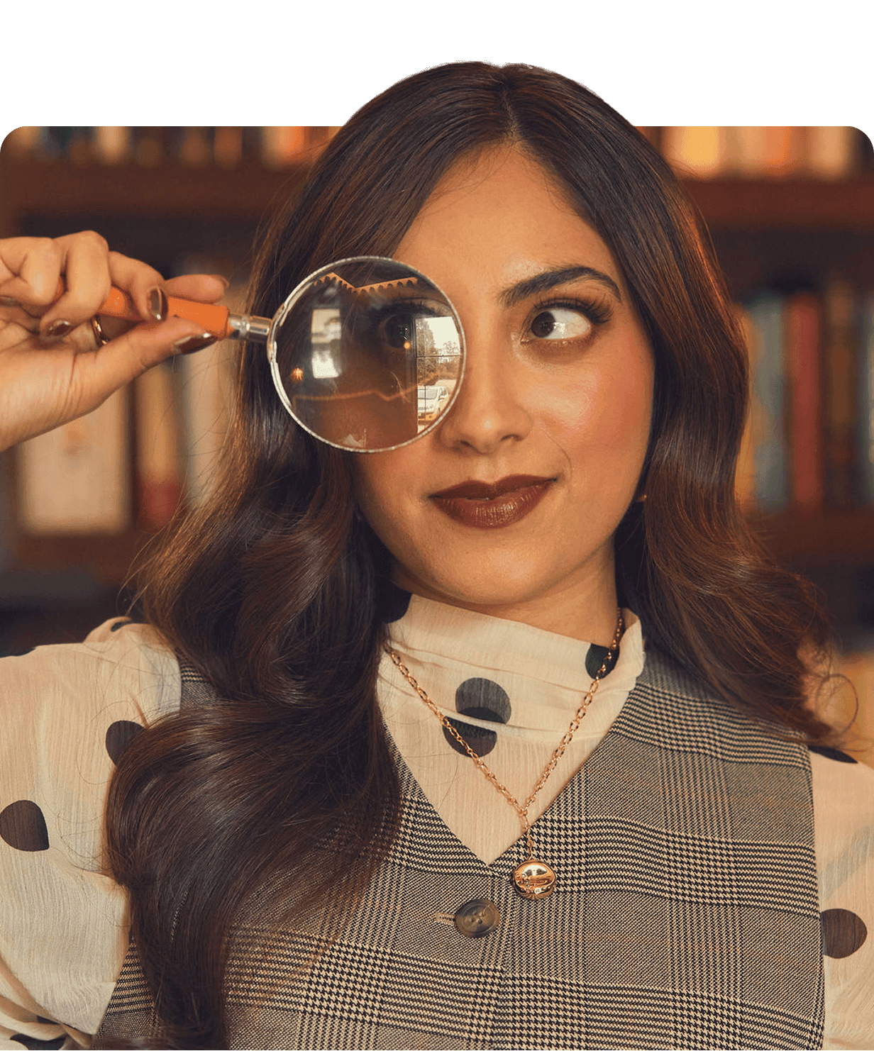 A woman with long dark hair holds a magnifying glass over one eye, standing in front of a blurred bookshelf. She wears a patterned top and layered necklaces.