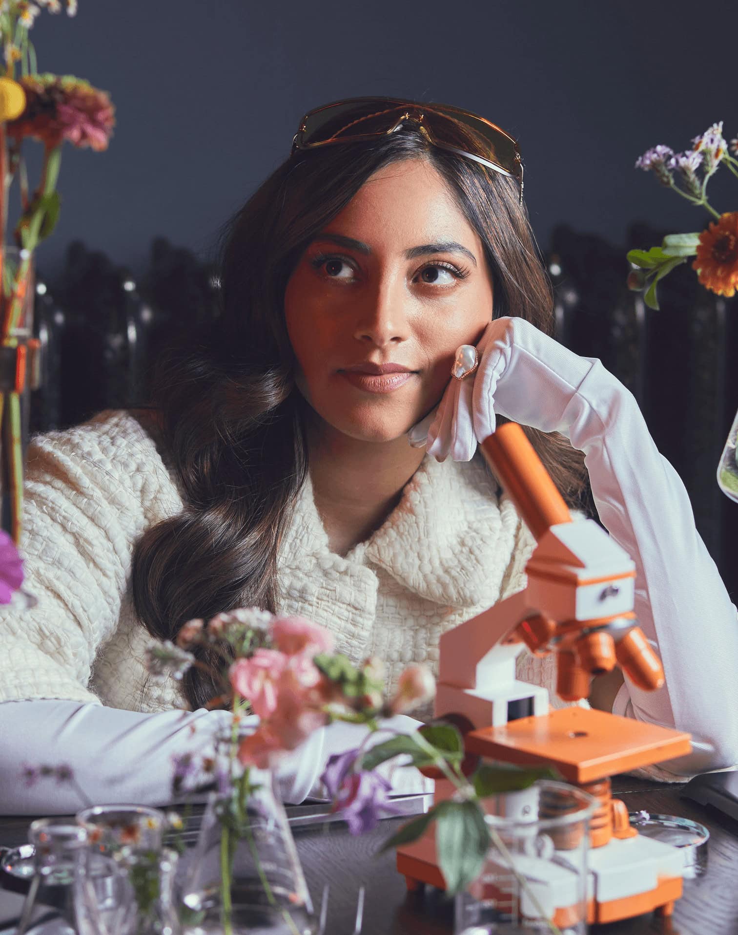 A woman wearing white gloves and a cream sweater sits at a table with flowers and an orange microscope, resting her chin on her hand and looking thoughtfully to the side.
