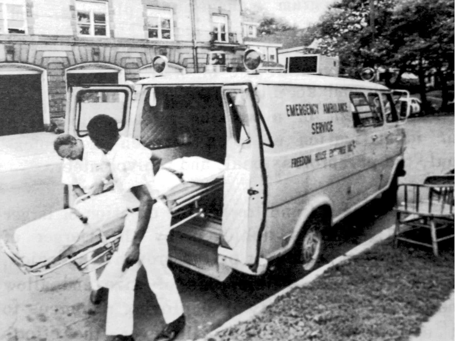 Two paramedics load a patient on a stretcher into an ambulance labeled "Emergency Ambulance Service" parked on a street outside a building.