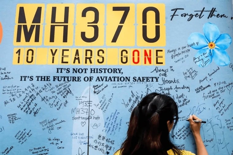 A person writes a message on a memorial wall for MH370's 10th anniversary, surrounded by handwritten notes and a sign reading "MH370 10 Years Gone.